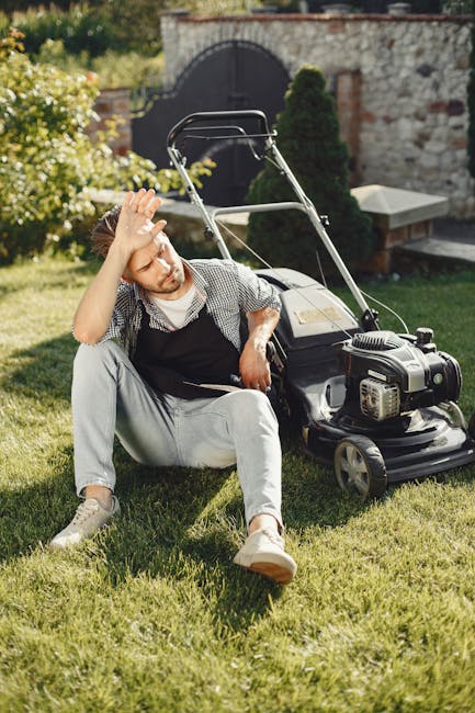 A young man with reddish hair and a beard, dressed in casual clothing including a checkered shirt and light-colored trousers, sits on a neatly mown lawn in a garden. Behind him, there is a black petro