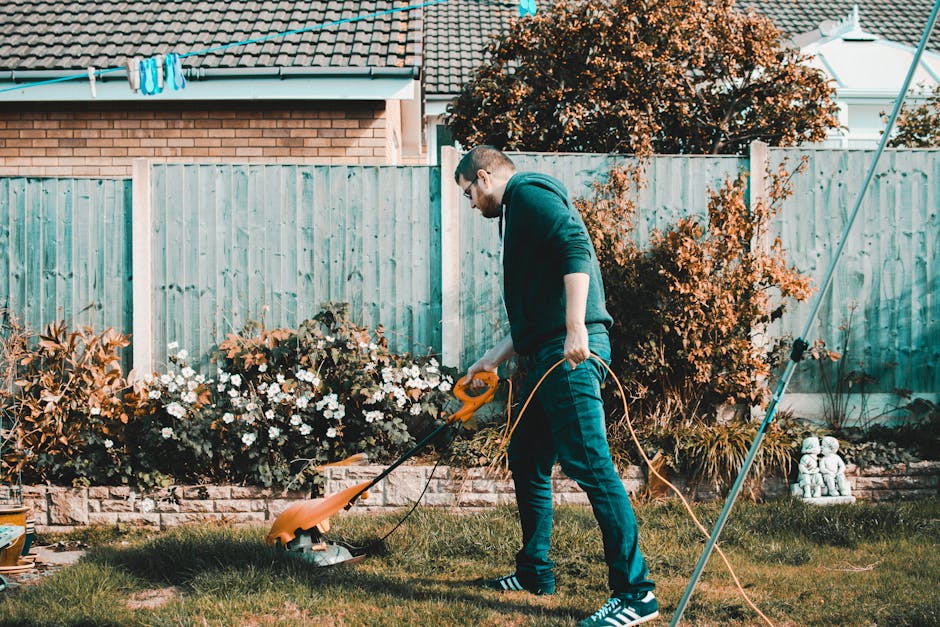 A young man with reddish hair and a beard, dressed in casual clothing including a checkered shirt and light-colored trousers, sits on a neatly mown lawn in a garden. Behind him, there is a black petro